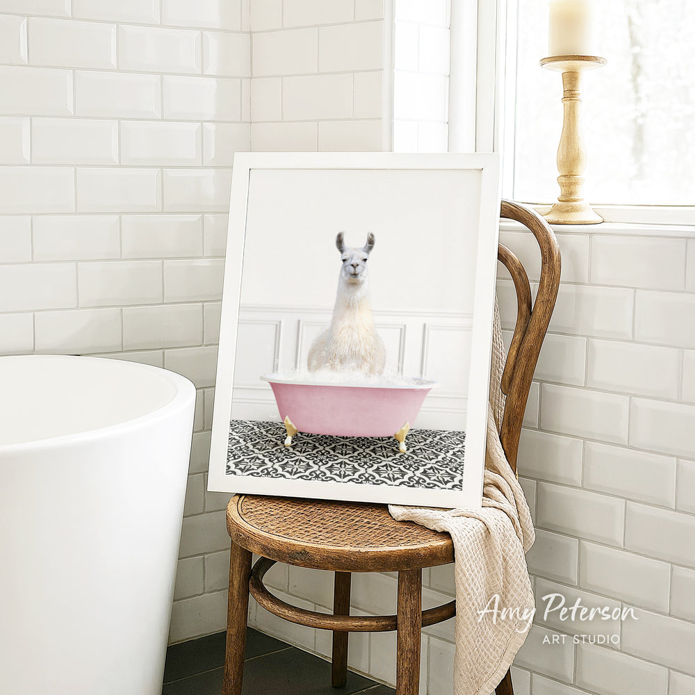 A framed image of a white llama sitting in a pink bathtub is displayed on a wooden chair in a bathroom setting.