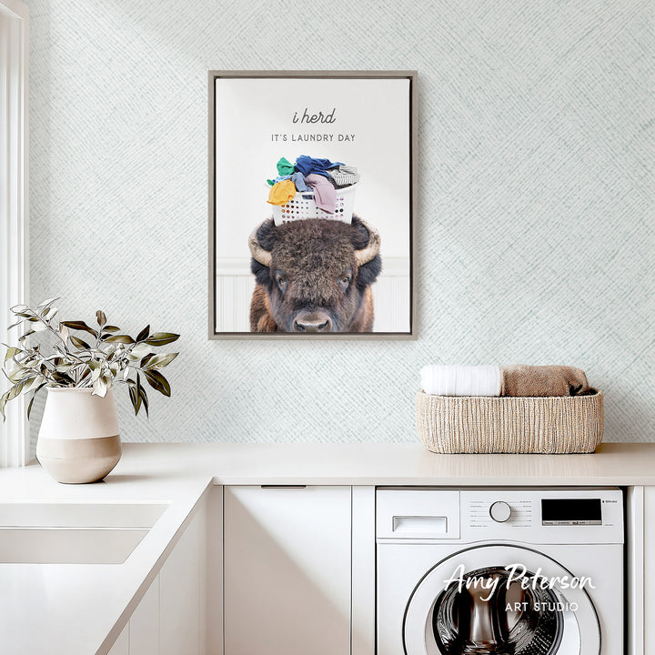 a framed poster of a buffalo with a laundry basket on its head, hanging on a wall above a white countertop with a potted plant and a basket of folded towels.