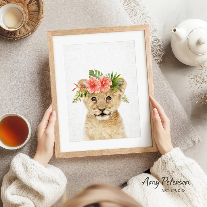 A person is holding a framed print of a lion wearing a floral crown, with a teapot and a cup of tea nearby.