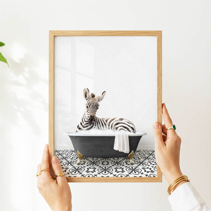 A person's hands are holding a picture frame with a framed image of a zebra sitting in a bathtub.