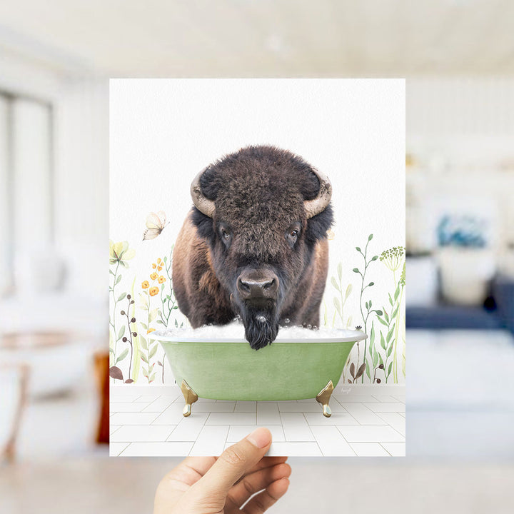 A hand holds up a framed image of a bison in a bathtub, surrounded by flowers and plants.