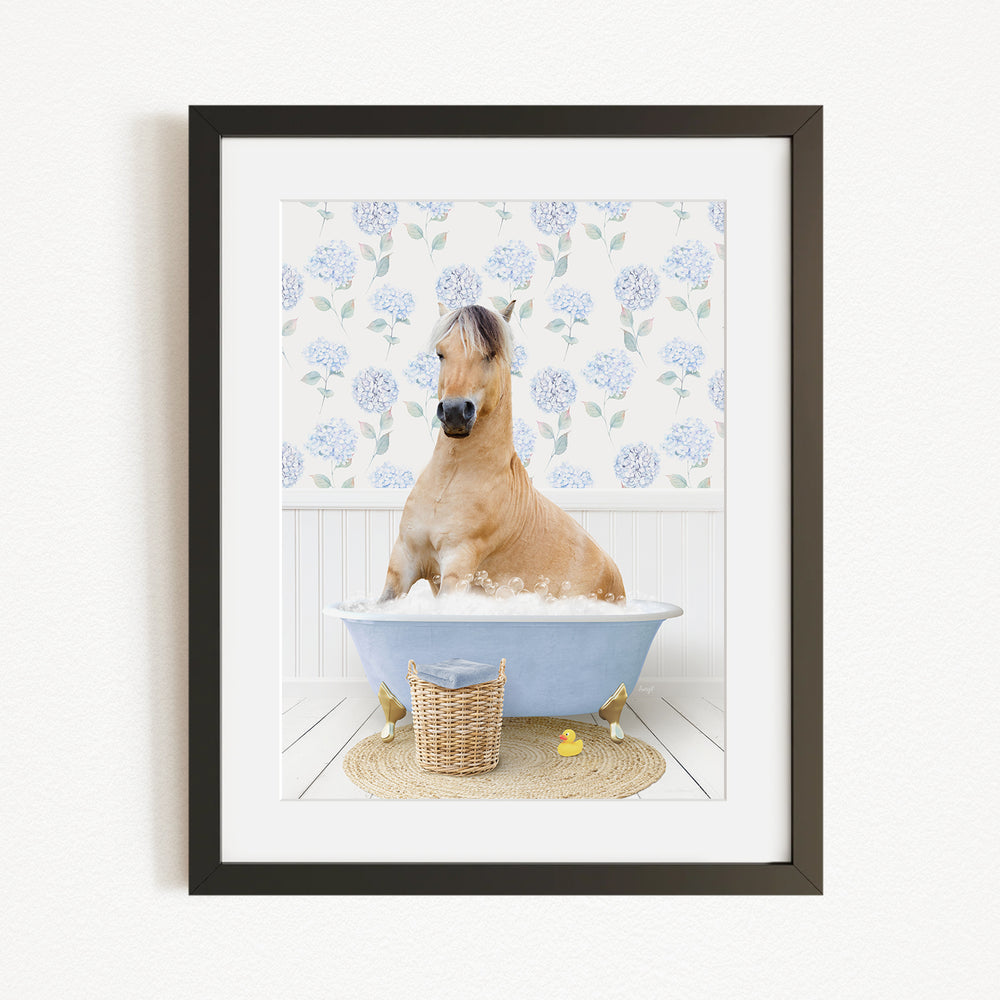 A light brown horse is sitting in a blue bathtub filled with bubbles, surrounded by a floral patterned wall and a basket of rubber ducks.