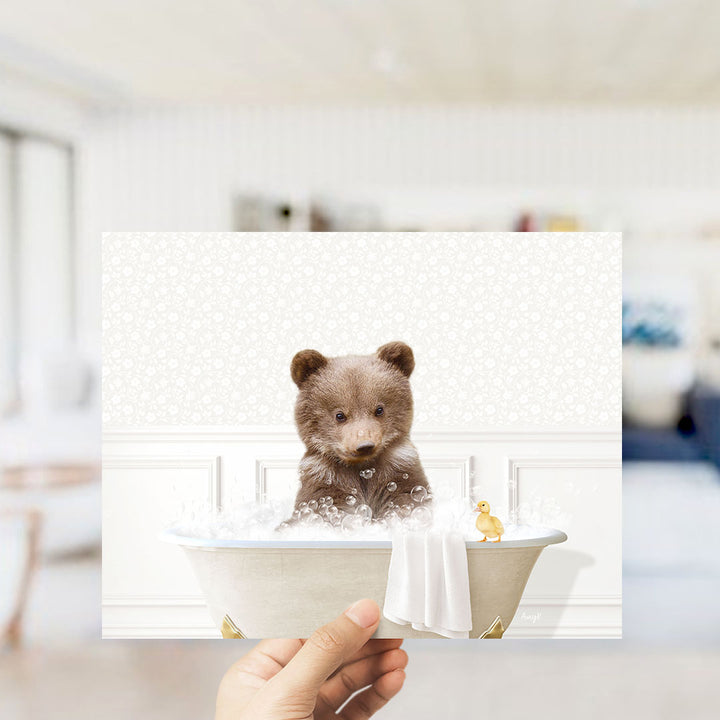 A hand holding a framed image of a brown bear sitting in a bathtub filled with bubbles, with a yellow rubber duck floating nearby.