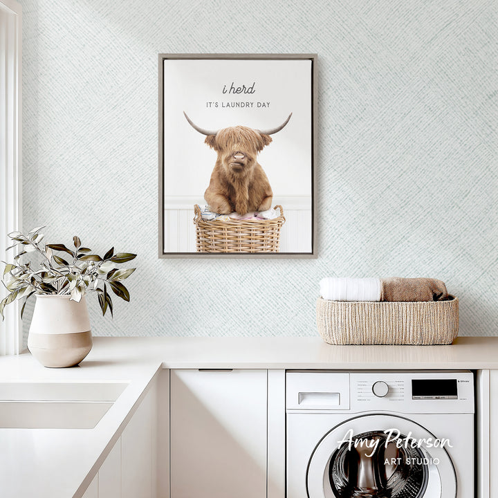 a framed poster of a Highland cow sitting in a wicker basket, with the caption "It's laundry day" above it. The poster is displayed on a wall above a white countertop, which has a potted plant and a basket of towels. A washing machine is also visible in the background.