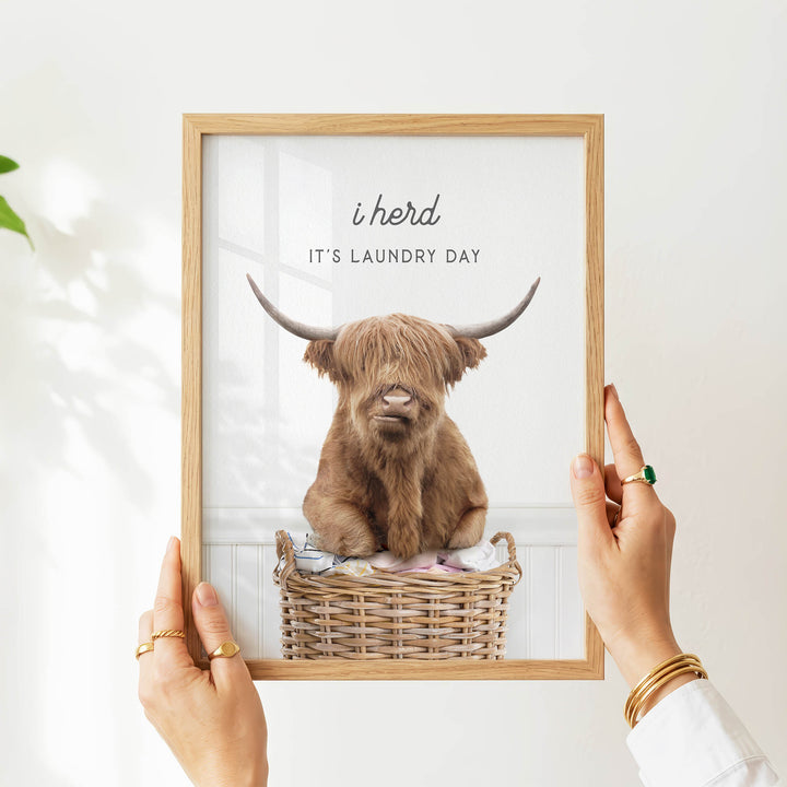 A person is holding up a framed picture of a brown cow with large horns, sitting in a wicker basket.