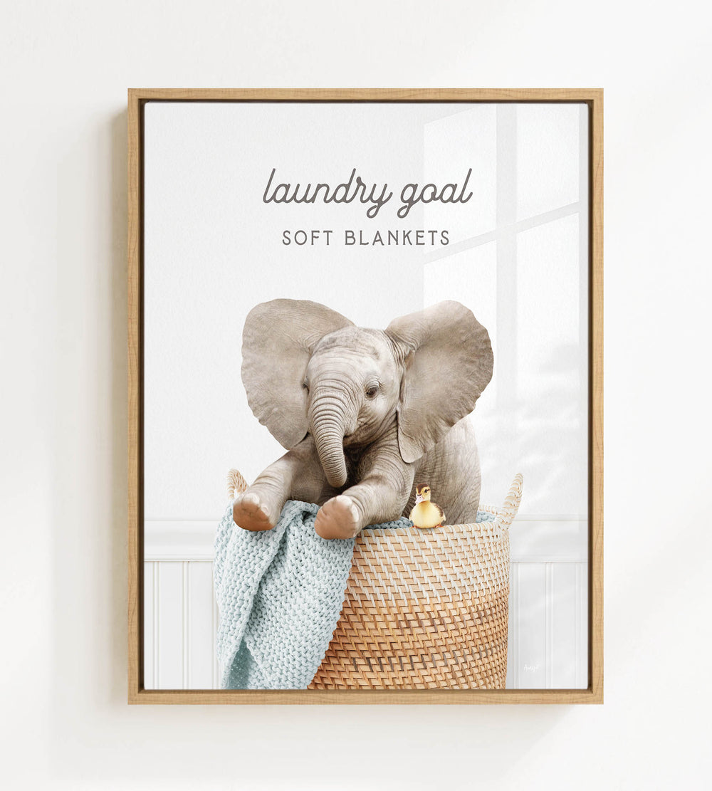 A framed image of an elephant sitting in a woven basket, with a small yellow bird perched on the edge of the basket.