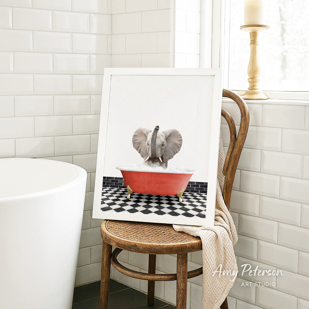 A framed image of a baby elephant sitting in a red bathtub, surrounded by a black and white checkered floor, is displayed on a wooden chair in a bathroom setting.