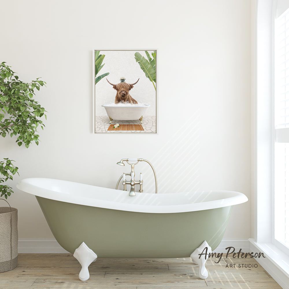A framed image of a Highland cow sitting in a bathtub is displayed on the wall above a green clawfoot bathtub in a bright, white-walled bathroom.