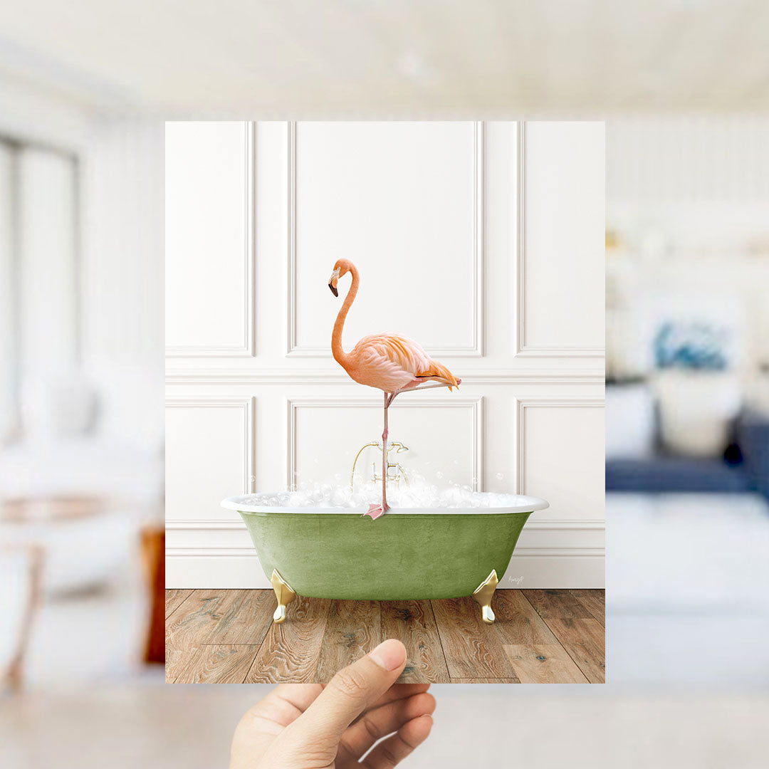 A hand holds up a photograph of a pink flamingo standing on a green bathtub filled with bubbles, set against a white wall with a wooden floor.