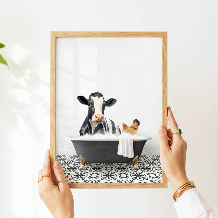 A framed image of a cow sitting in a bathtub, with a chicken nearby, is being held up by a person's hands.