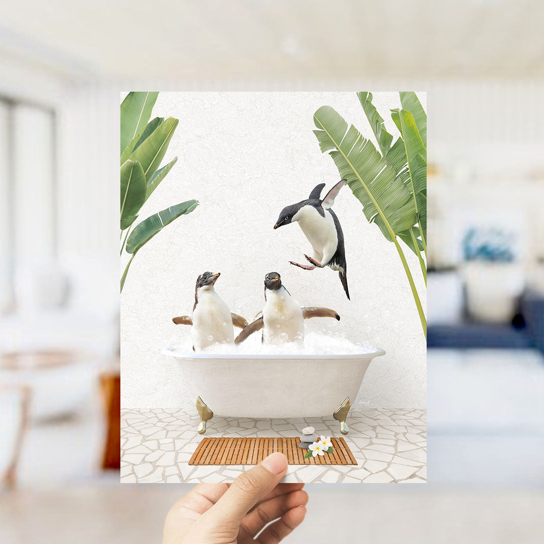 A hand holds up a framed image depicting two penguins sitting in a bathtub, surrounded by tropical plants.