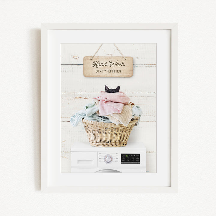 A framed image of a cat sitting in a basket filled with laundry, with a sign above that says "Hand Wash Dirty Kittens".