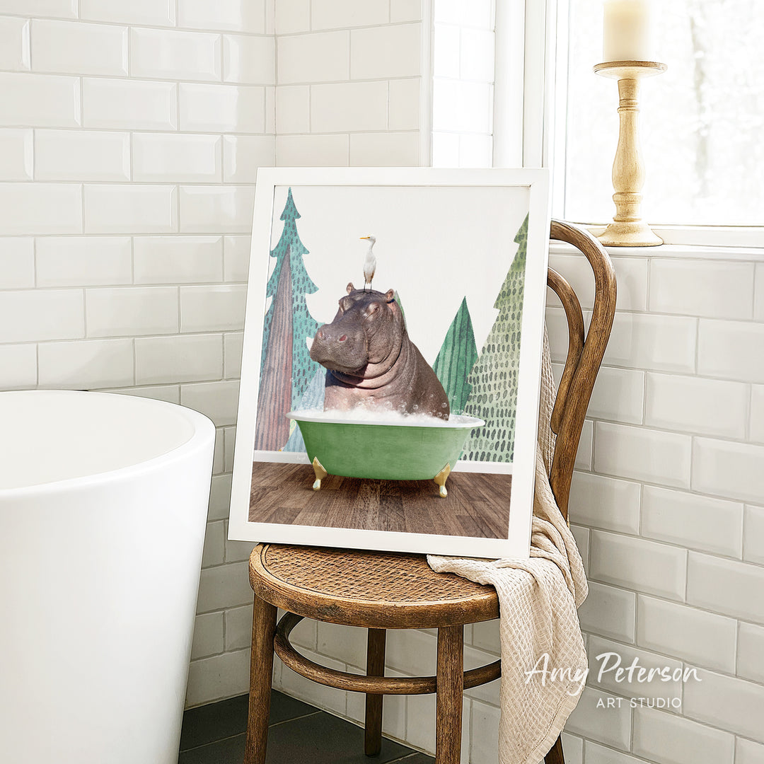A framed image of a hippopotamus sitting in a green bathtub, surrounded by trees, is displayed on a wooden chair in a bathroom setting.