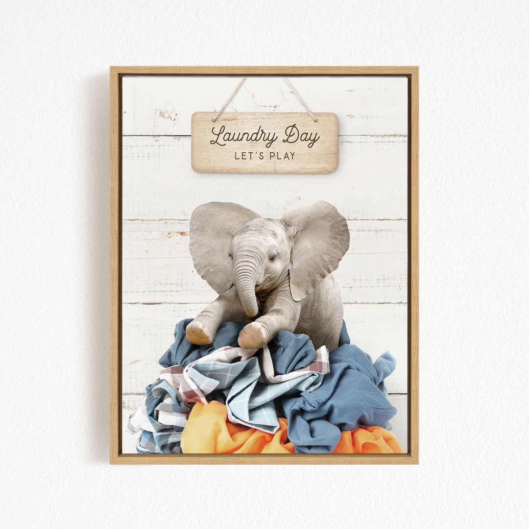 A framed image of a stuffed elephant sitting atop a pile of laundry, with a wooden sign above that reads "Laundry Day Let's Play".