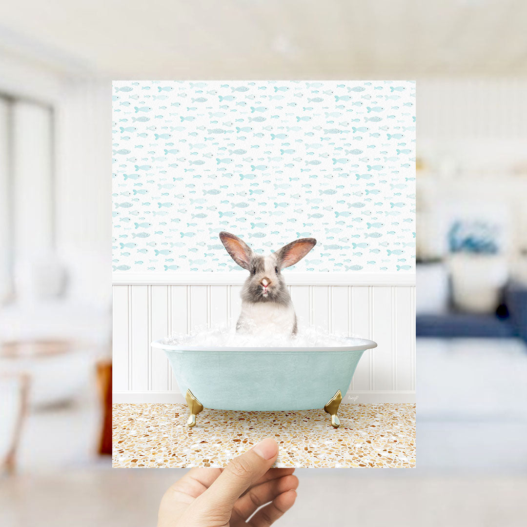 A hand holds up a collage of images featuring a rabbit sitting in a bathtub filled with bubbles, surrounded by a pattern of fish.