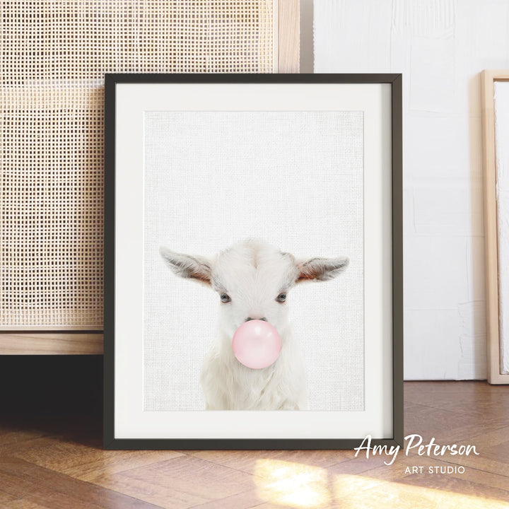A framed image of a white goat blowing a bubble with pink gum, set against a white background.