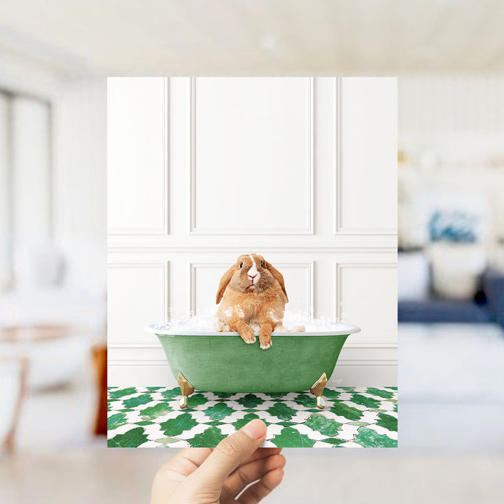 A hand holds up a framed image of a brown rabbit sitting in a green bathtub filled with bubbles, surrounded by a green and white patterned floor.