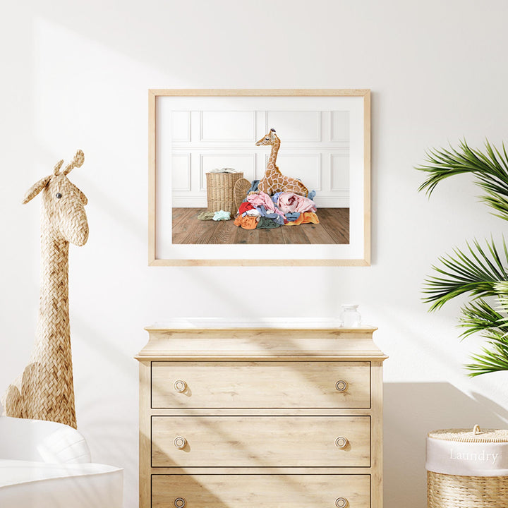 a giraffe sitting on a pile of clothes in a room with a wooden dresser and a potted plant.