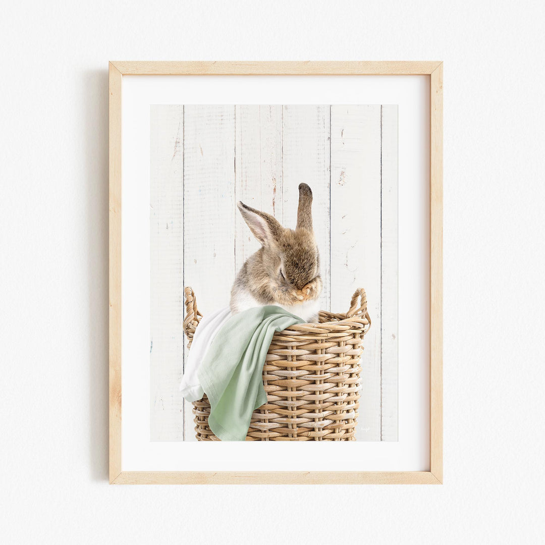 A small, fluffy rabbit sits in a wicker basket, with its head resting on a green blanket.