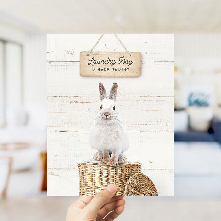 A hand is holding up a card with a white rabbit sitting on a wicker basket, with a wooden sign above that says "Laundry Day is Hare Raising".