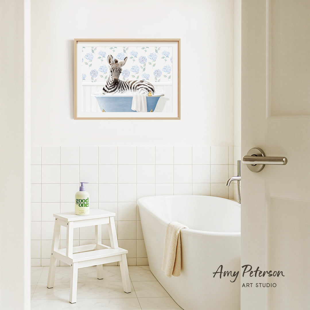 A framed painting of a zebra sitting in a bathtub is hanging on a white tiled wall above a white bathtub in a bathroom.
