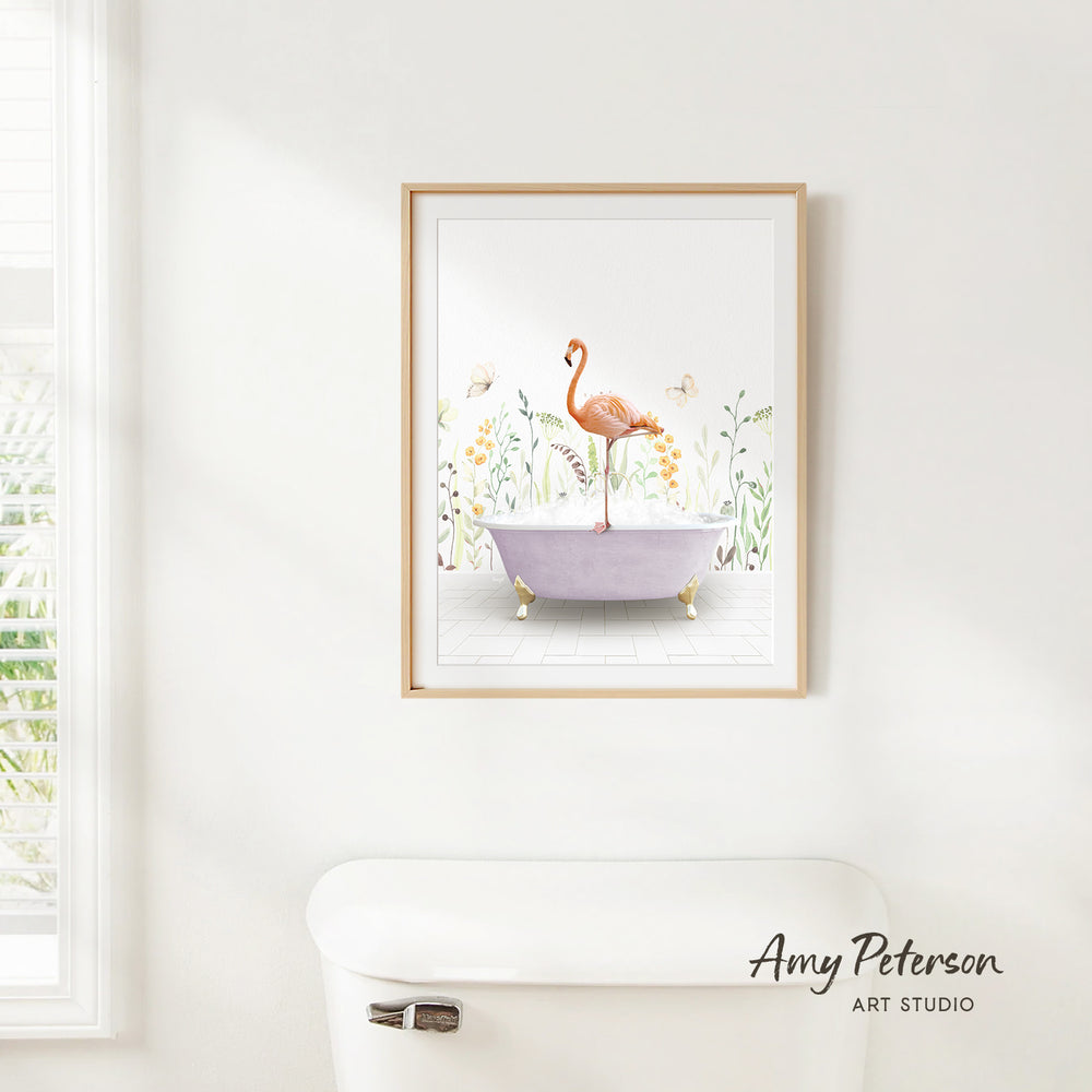 A framed illustration of a pink flamingo standing on a purple bathtub, surrounded by flowers and greenery, is hanging on a white wall.