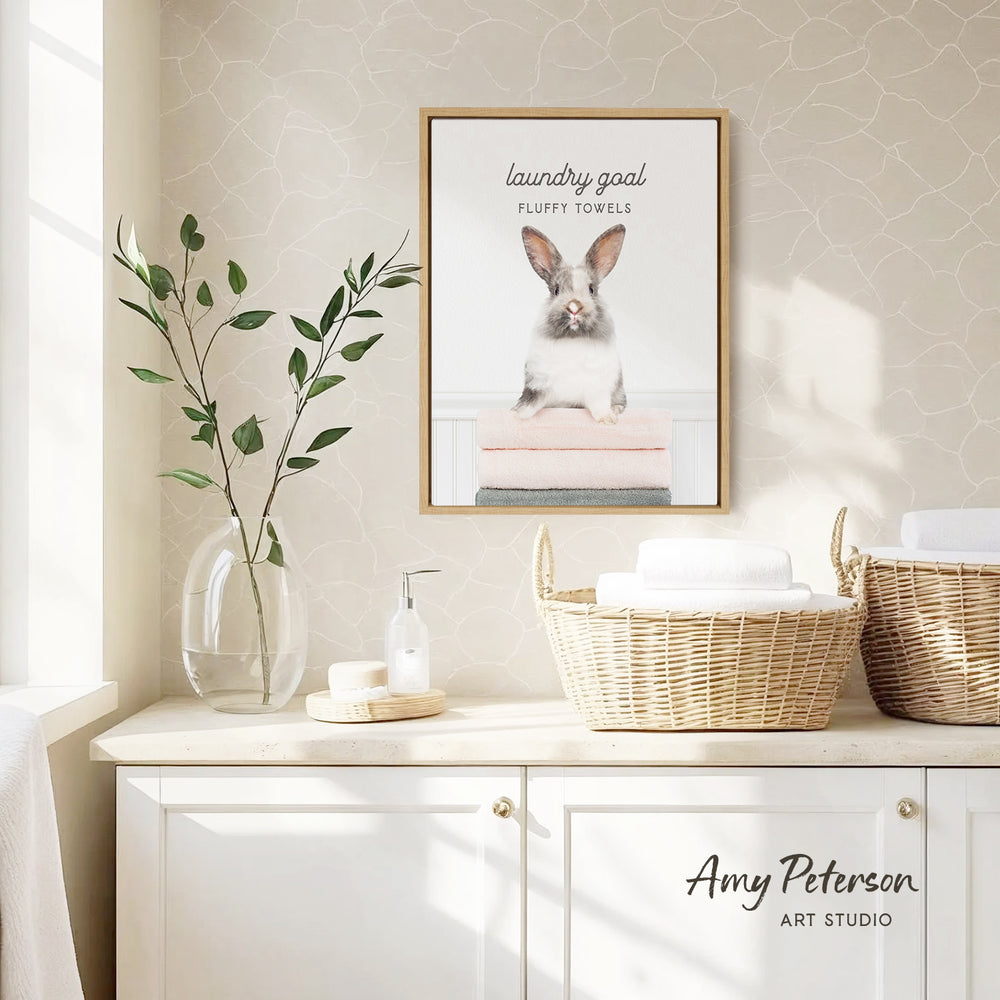 a framed poster of a rabbit sitting on a pile of fluffy towels, which is hanging on a wall above a white cabinet. The cabinet has two woven baskets on top of it, and there is a vase with a plant on the counter. The overall scene has a clean and organized appearance.