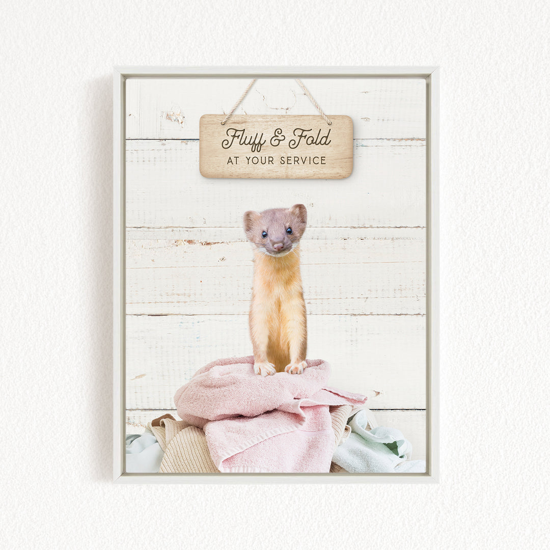 A small brown and white ferret sitting on a pile of clothes, with a wooden sign above it that says "Fluff & Fold at your service".