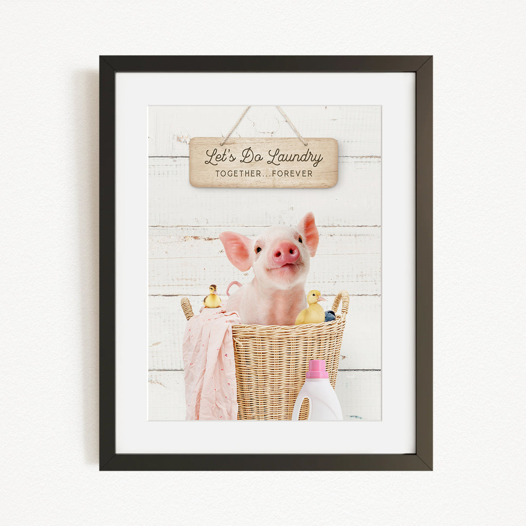 A framed image of a pig sitting in a wicker basket filled with rubber ducks, with a sign above that reads "Let's Do Laundry Together Forever".