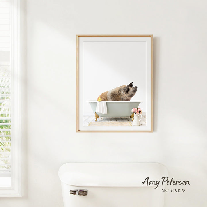 A framed image of a pig sitting in a bathtub, with a vase of flowers and a white toilet visible in the background.