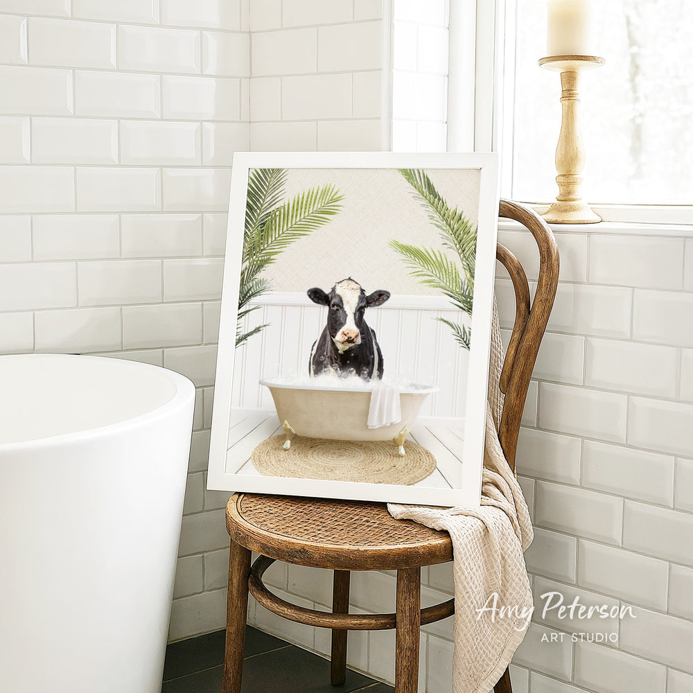 A framed image of a cow sitting in a bathtub, surrounded by palm leaves, is displayed on a wooden chair in a bathroom setting.