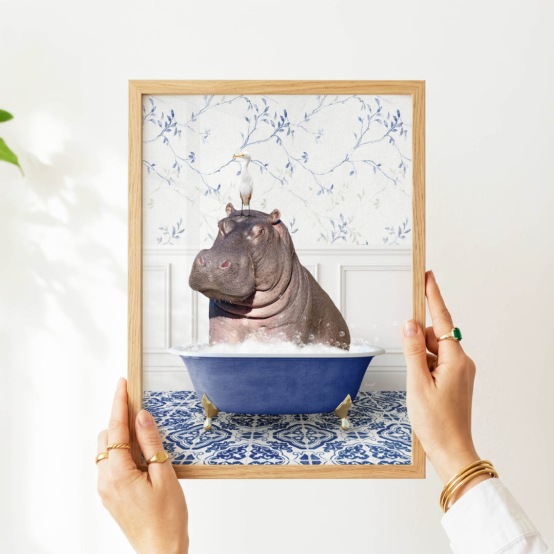 A person's hands are holding a framed image of a hippopotamus sitting in a blue bathtub filled with water, surrounded by a blue and white floral pattern.