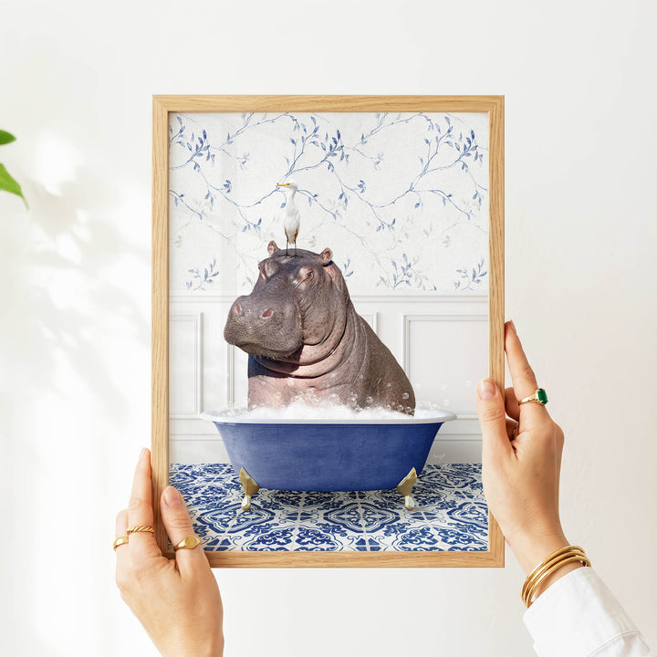 A person's hands are holding a framed image of a hippopotamus sitting in a blue bathtub filled with water, surrounded by a blue and white floral pattern.