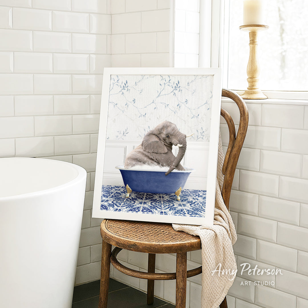 A framed image of a gray elephant sitting in a blue bathtub, surrounded by a blue and white floral pattern, is displayed on a wooden chair in a bathroom setting.