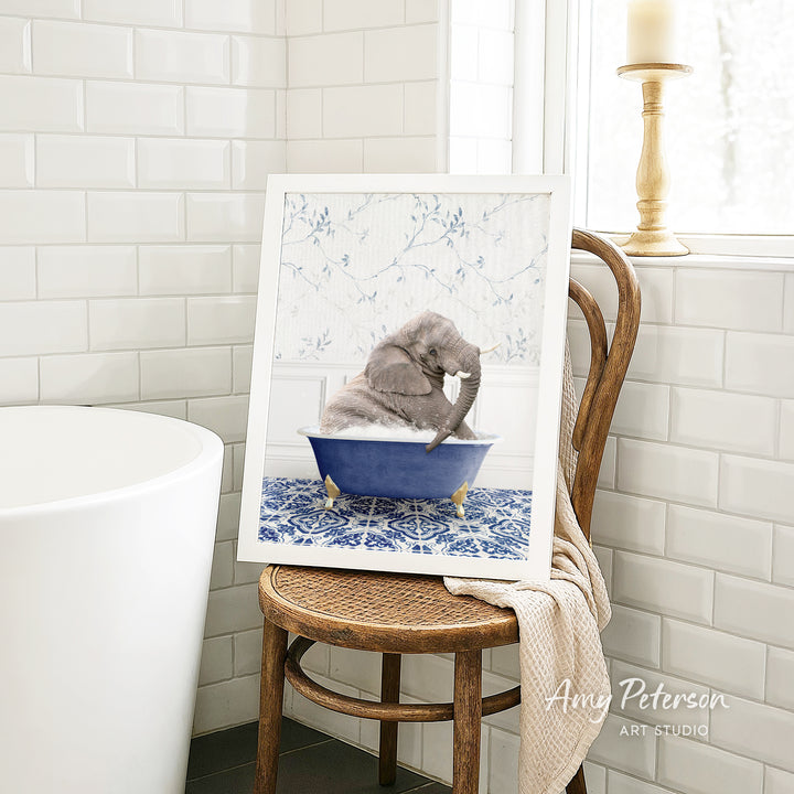A framed image of a gray elephant sitting in a blue bathtub, surrounded by a blue and white floral pattern, is displayed on a wooden chair in a bathroom setting.