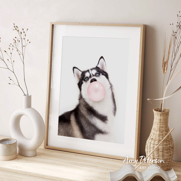 A framed photograph of a husky dog blowing a bubble with a pink bubble gum bubble, accompanied by two vases and a woven basket on a wooden surface.
