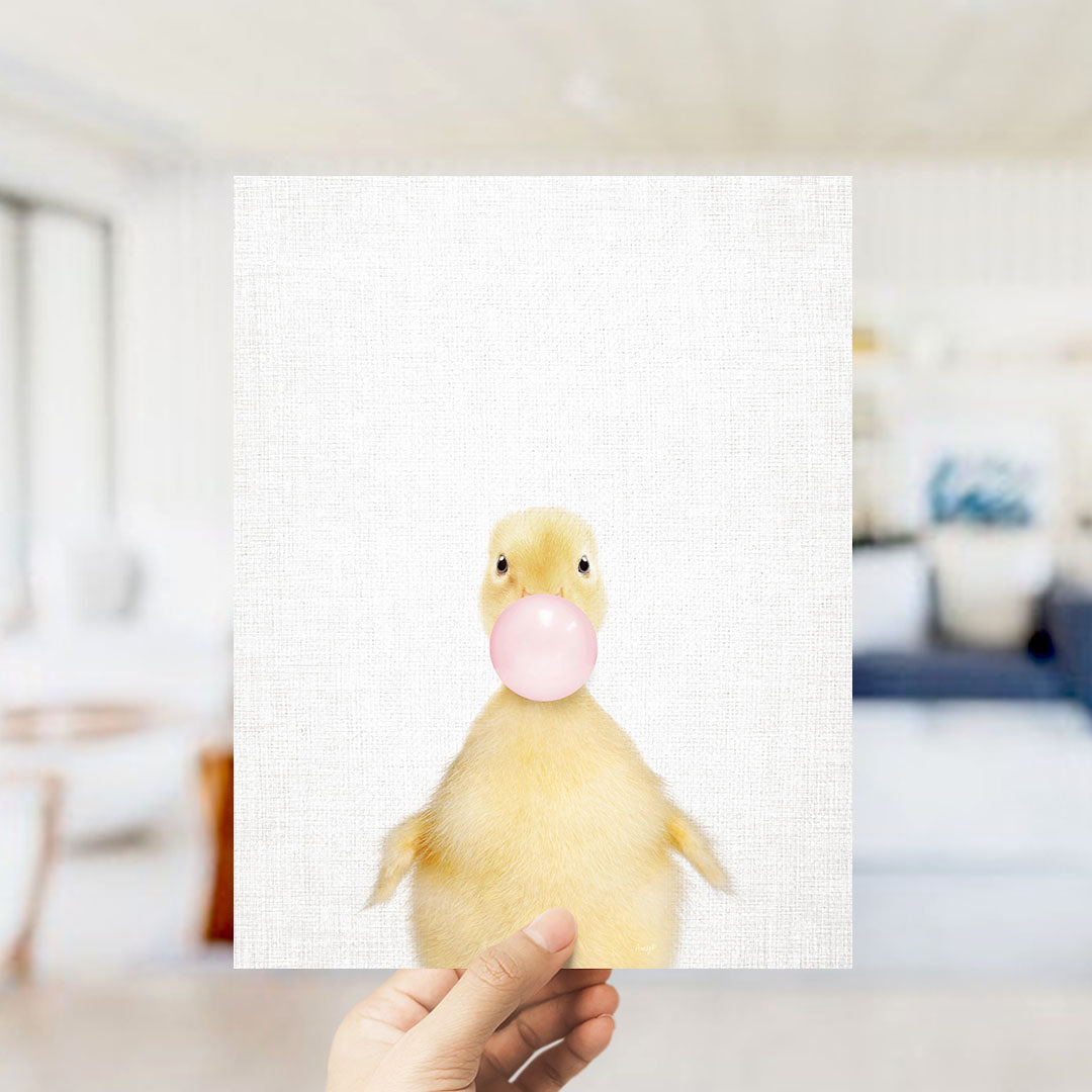 A hand is holding up a white square with a cute yellow duck blowing a bubble with a pink bubble gum bubble.