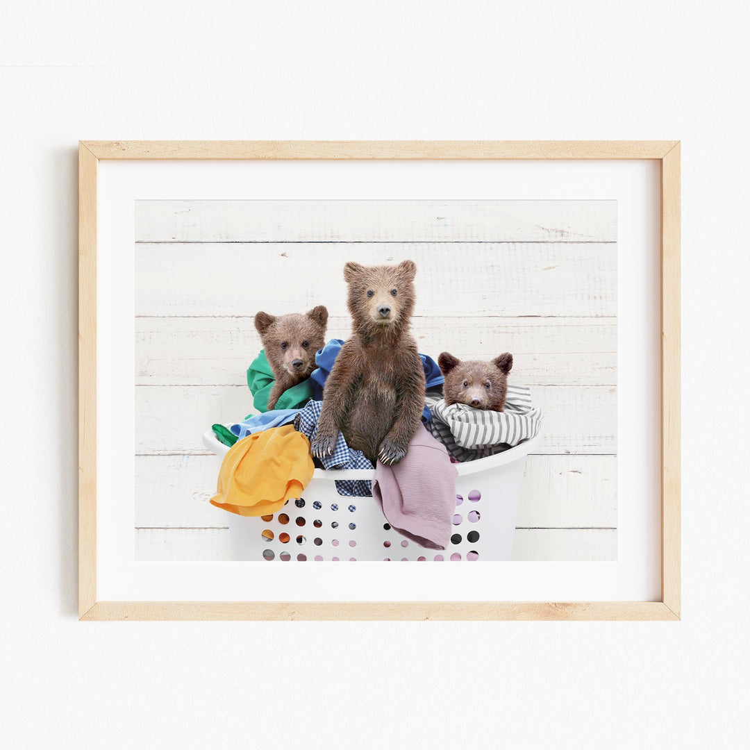 A framed image depicting three brown bears sitting in a laundry basket filled with clothes.