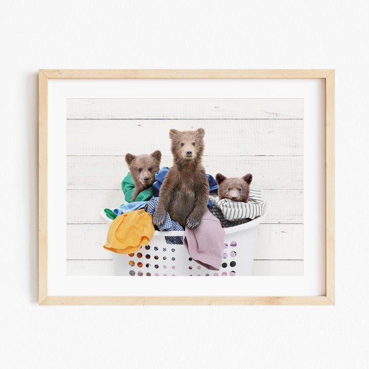 A framed image depicting three brown bears sitting in a laundry basket filled with clothes.