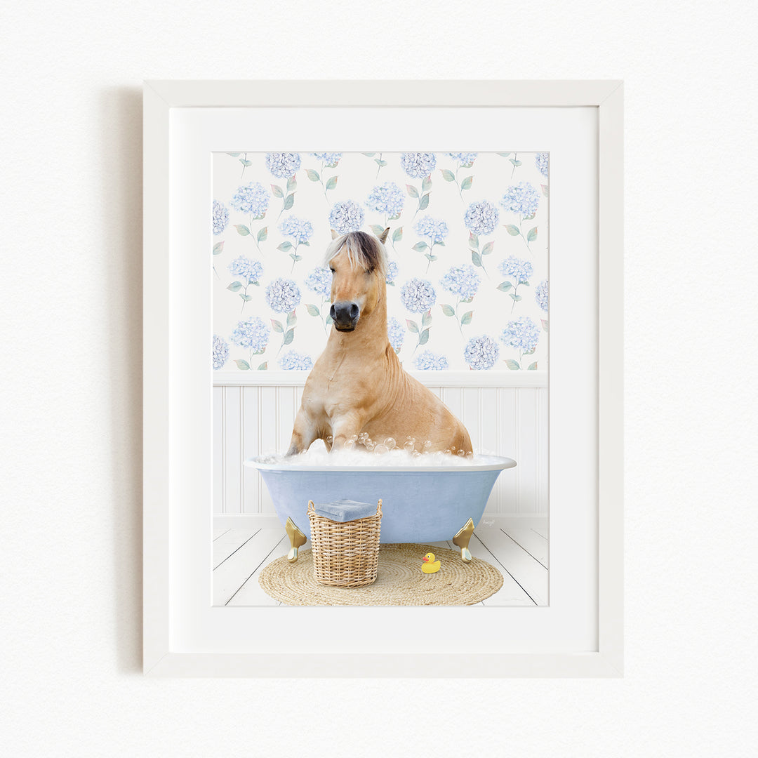 A light brown horse is sitting in a blue bathtub filled with water, surrounded by a white floral patterned wall and a basket of rubber ducks.