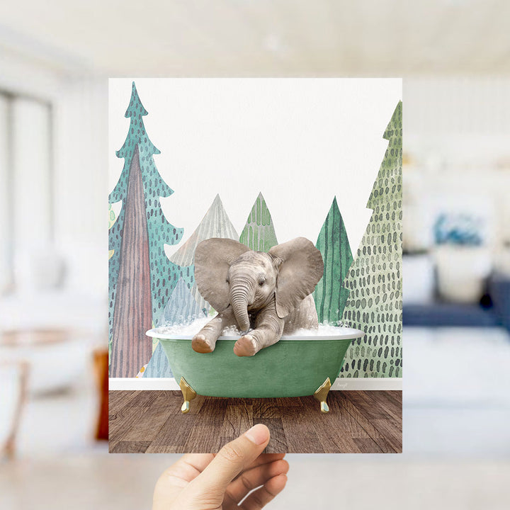 A hand holds up a framed image depicting a baby elephant sitting in a green bathtub, surrounded by a forest scene with trees and a blue sky.