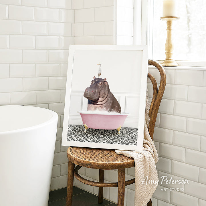 A framed image of a hippopotamus sitting in a pink bathtub, with a bird perched on its head, is displayed on a wooden chair in a bathroom setting.