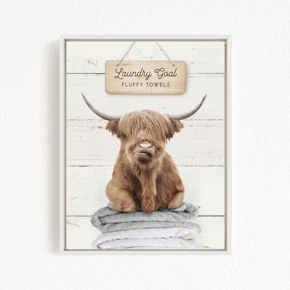 A framed image of a brown cow with long horns sitting on a stack of fluffy towels, with a sign above that says "Laundry Goal Fluffy Towels".