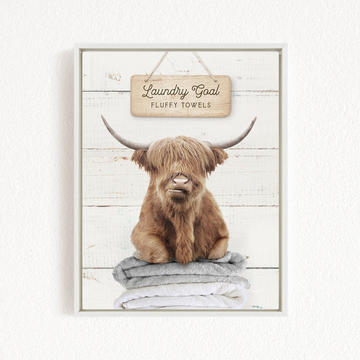 A framed image of a brown cow with long horns sitting on a stack of fluffy towels, with a sign above that says "Laundry Goal Fluffy Towels".