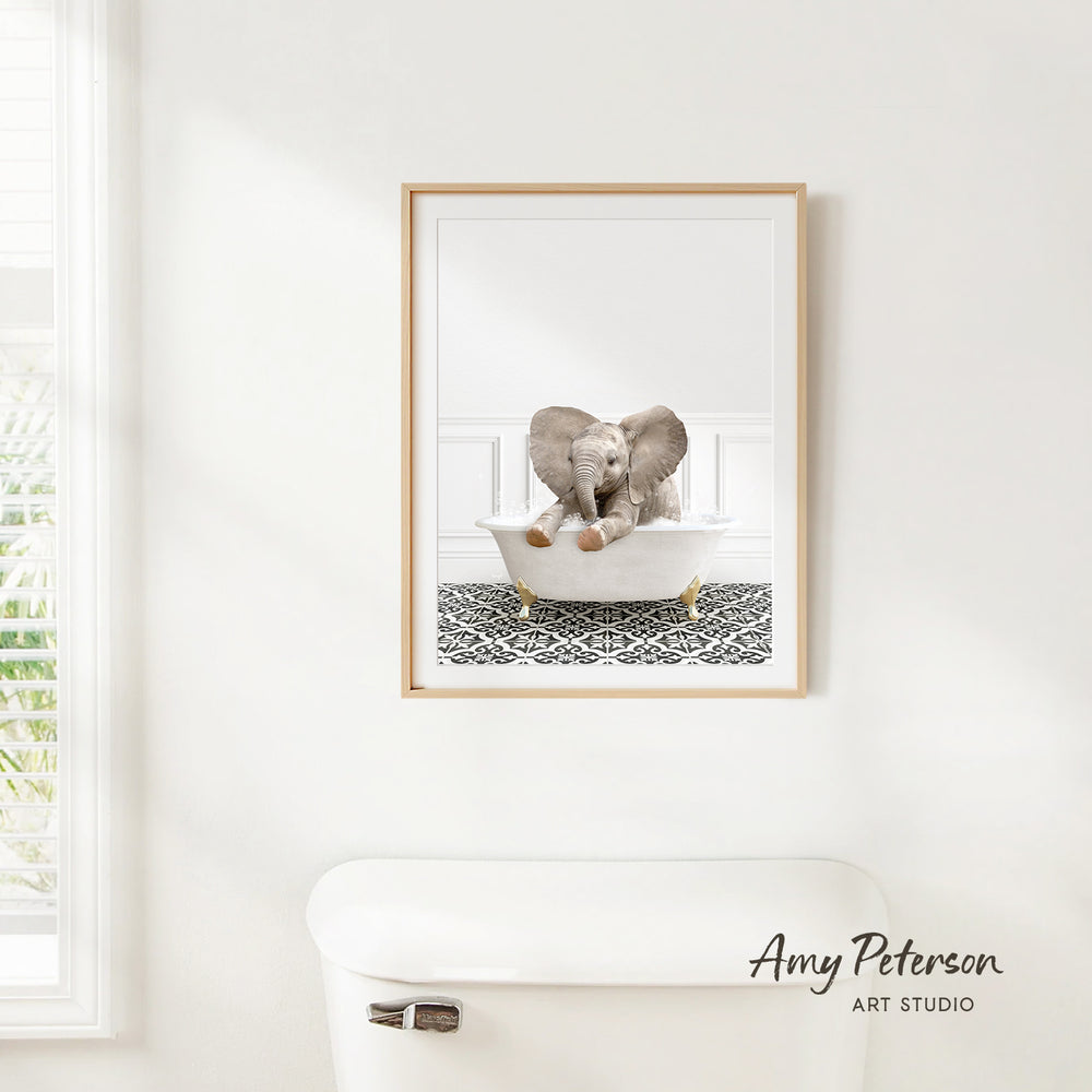 A framed image of an elephant sitting in a bathtub, with a patterned tile floor beneath it, is hanging on a white wall.