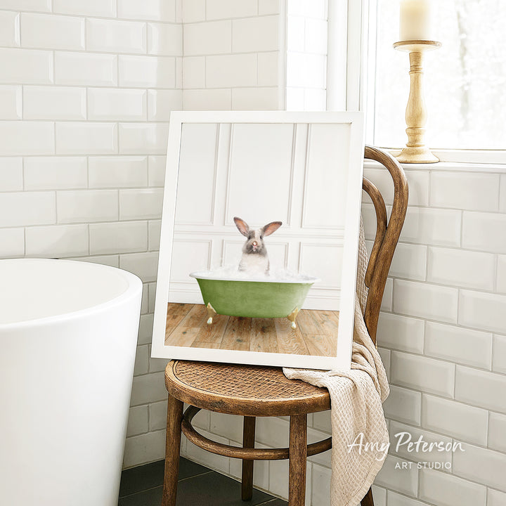 A framed image of a rabbit sitting in a green bathtub, with a wooden chair and a white tiled wall in the background.