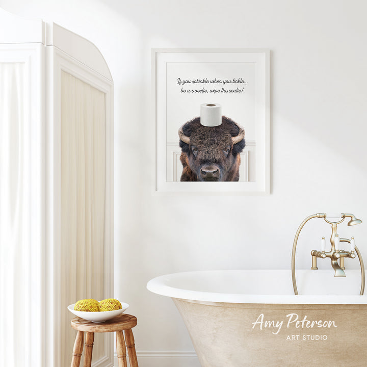 a bathroom with a framed picture of a buffalo on a toilet paper roll, a wooden stool with a bowl of fruit, and a bathtub with a faucet.