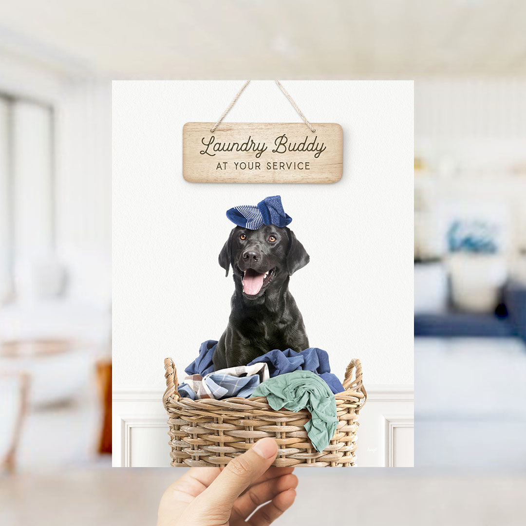 A black dog wearing a blue hat sits in a wicker basket filled with clothes, with a wooden sign above that reads "Laundry Buddy at your service".