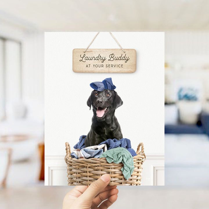 A black dog wearing a blue hat sits in a wicker basket filled with clothes, with a wooden sign above that reads "Laundry Buddy at your service".