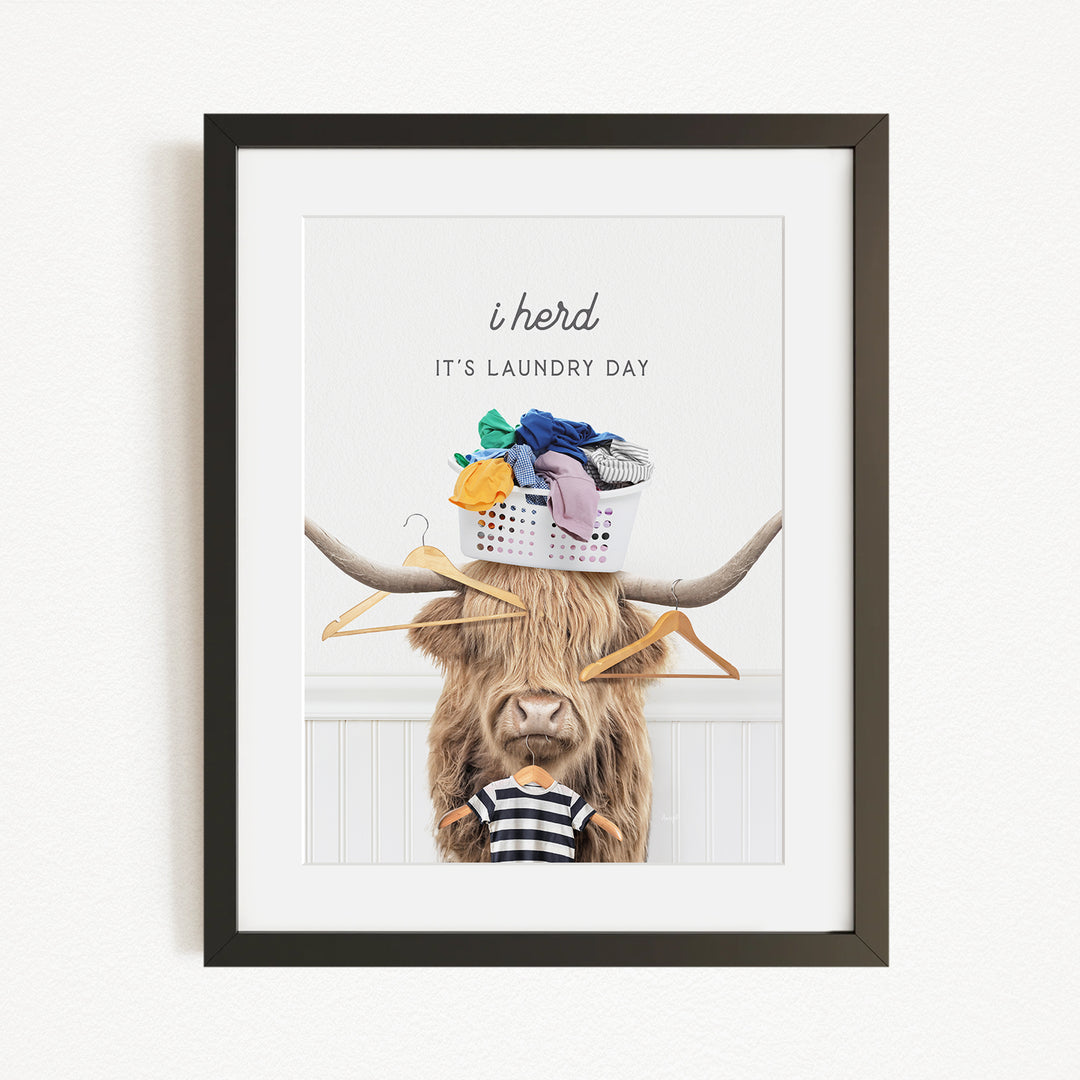 A framed image depicting a cow with a basket of laundry on its head, accompanied by the text "It's laundry day" and the phrase "i herd"